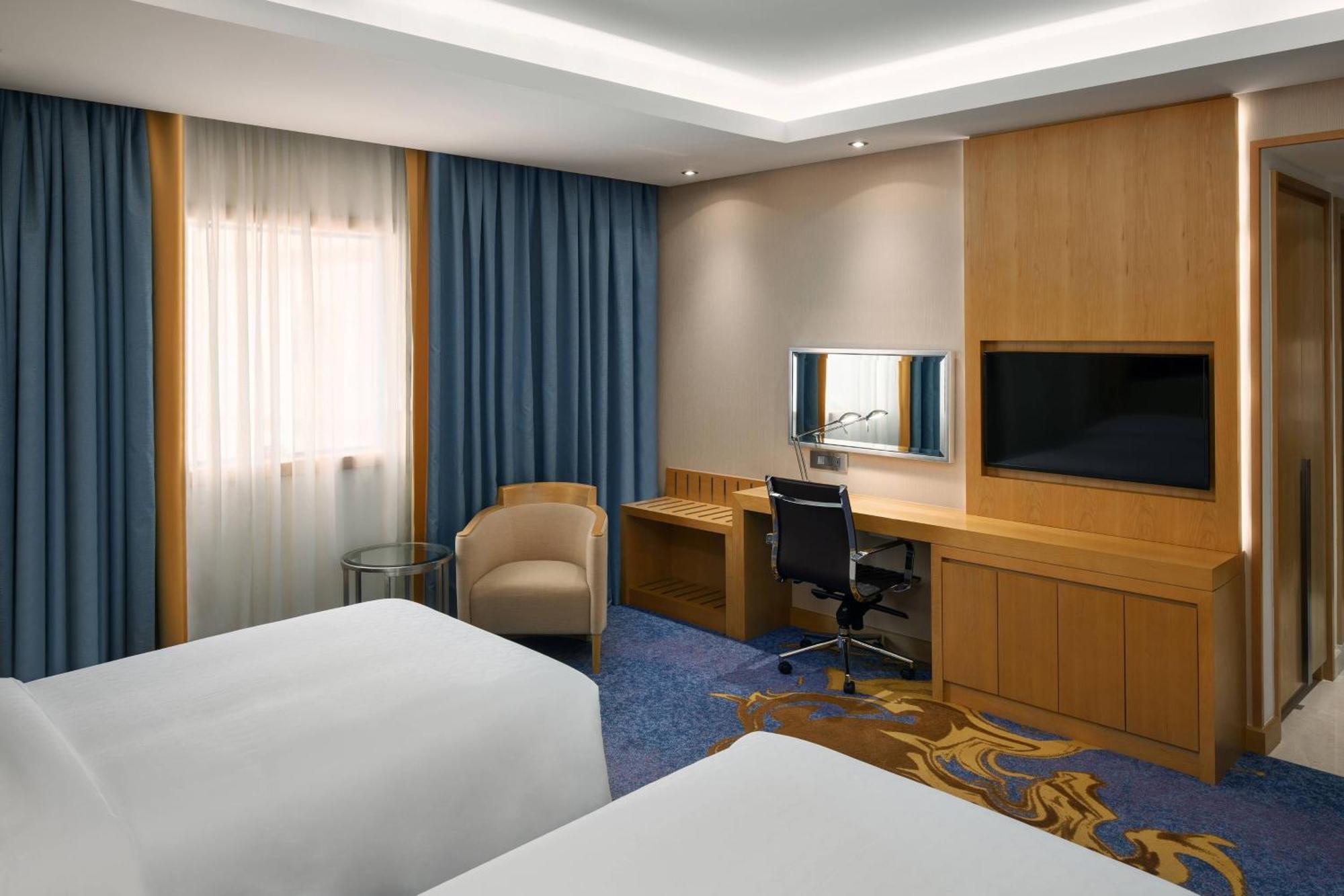 Sheraton Makkah Jabal Al Kaaba Hotel Μέκκα Εξωτερικό φωτογραφία The photo shows a hotel room featuring two beds with white linens. There are large windows covered with blue curtains that allow natural light to enter. On one side of the room, there is a wooden workstation with a chair and a mirror, along with a fl