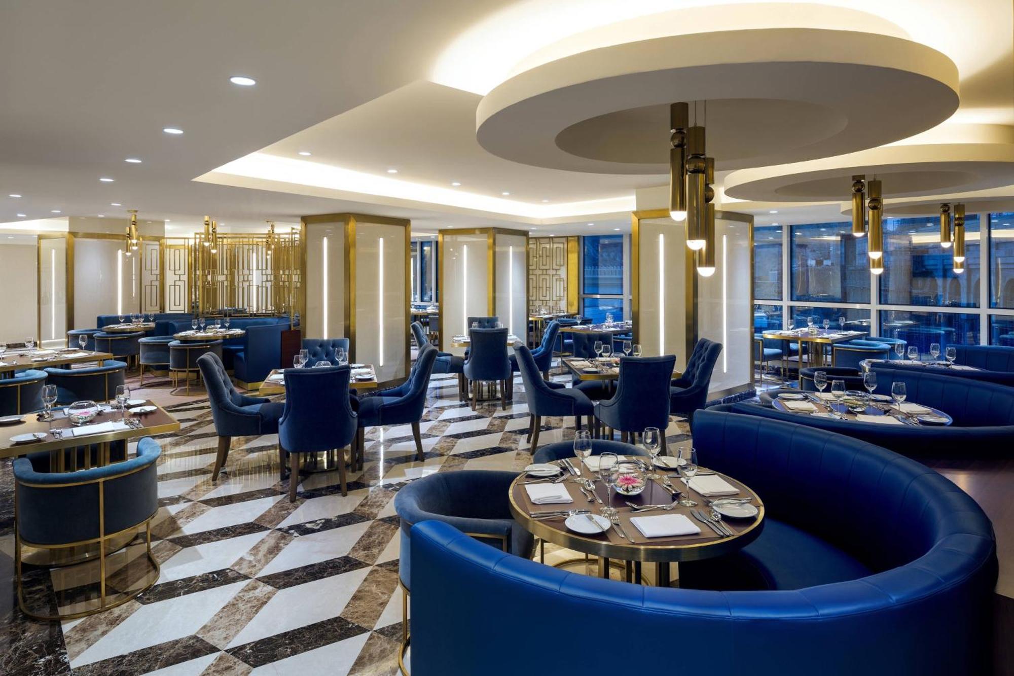 Sheraton Makkah Jabal Al Kaaba Hotel Μέκκα Εξωτερικό φωτογραφία The photo shows the interior of a modern restaurant. The design features elegant curved blue seating arranged around round tables with plates and glassware set for dining. The flooring is a striking marble pattern, adding to the upscale ambiance. The